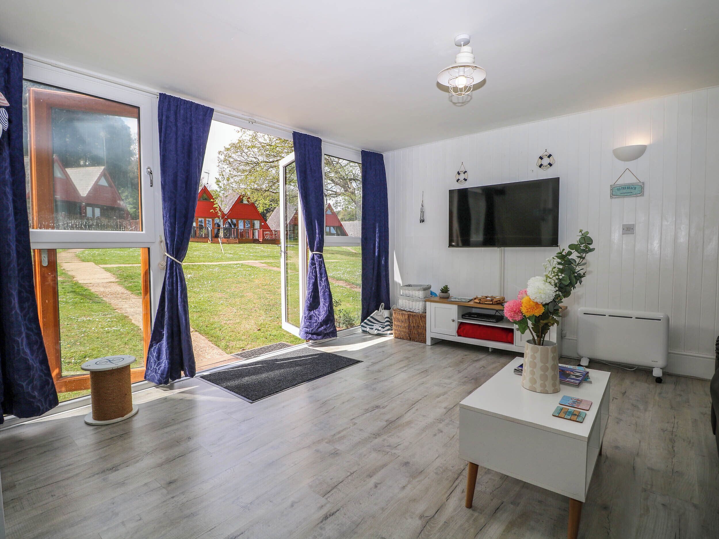Uma sala de estar moderna com uma televisão grande, uma mesa de centro branca e vista para uma casa com telhado vermelho através da porta de vidro.