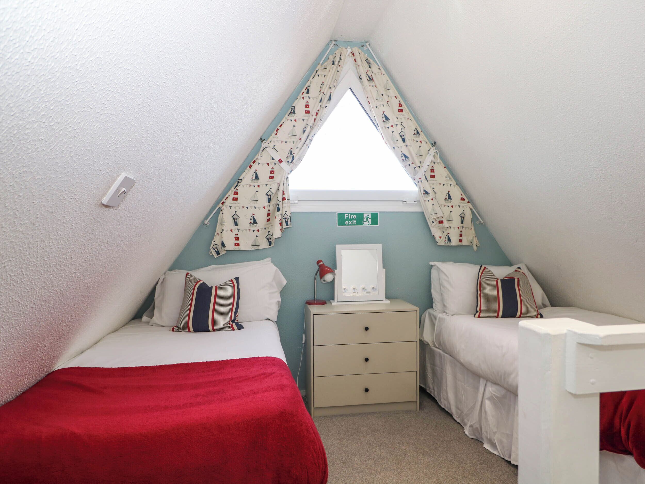 A small, triangular room with two beds, a dresser, and a window with a nautical-themed curtain. A fire exit sign is visible on the wall.
