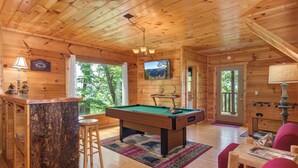 A wooden cabin interior with a pool table, bar area, and seating.