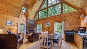 A cozy cabin interior with a fireplace, wooden walls, a dining area, and a kitchen.