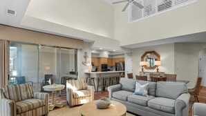 A spacious living room with a grey sofa, striped armchairs, and a round coffee table. A kitchen is visible in the background with modern appliances. - Lake View Villa at Hidden Dunes! (Miramar Beach)