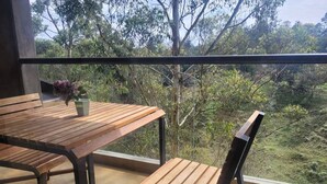 A balcony with a wooden table and chairs, a potted plant, and a view of trees and greenery.