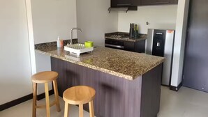 A modern kitchen with a granite countertop, two wooden stools, a built-in island, stainless steel appliances, and dark cabinetry.