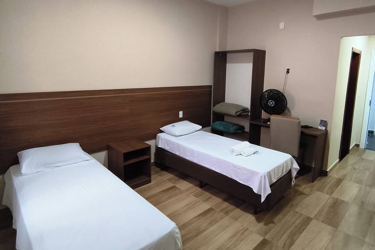 A hotel room with two single beds, a wooden headboard, a small desk with a chair, a mirror, a round table with a chair, and a door leading to another room.