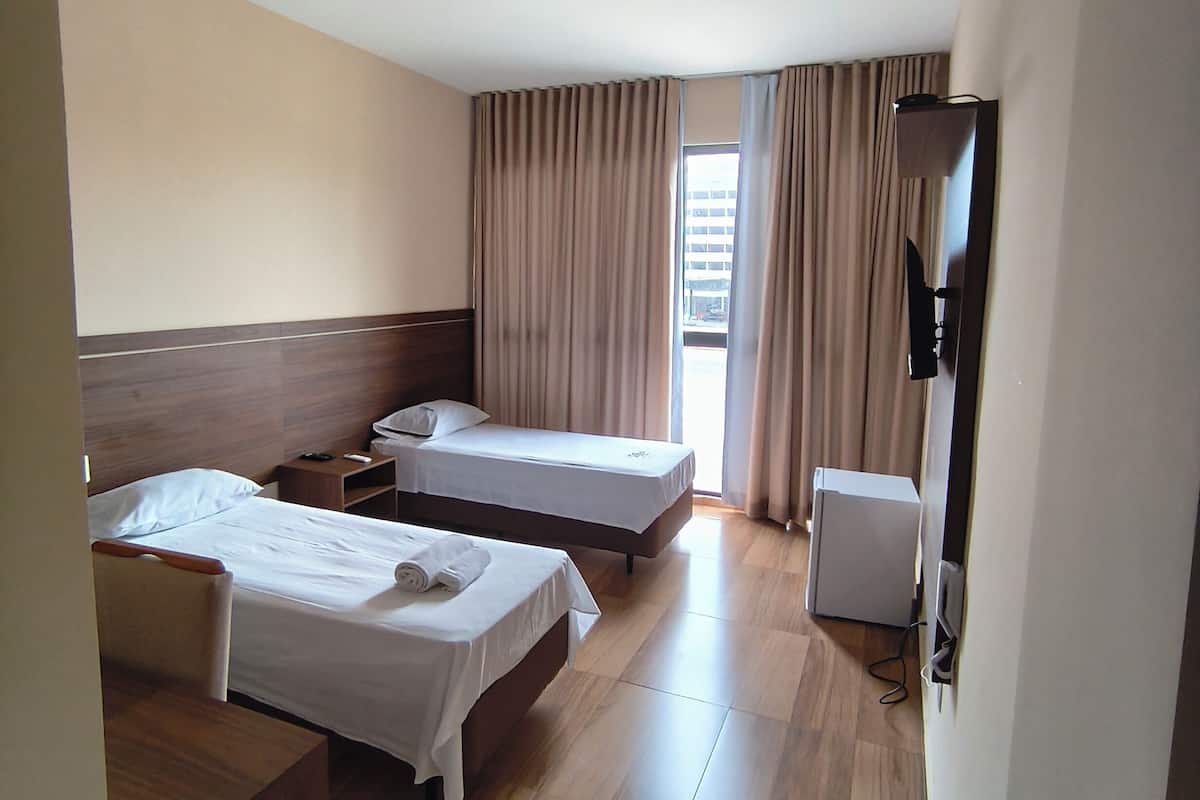 A hotel room with two beds, a desk, a chair, a bedside table, a lamp, a window with curtains, and a flat-screen TV.