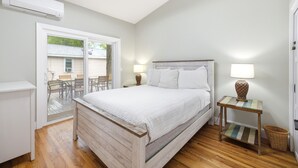 A bedroom with a bed, two bedside lamps, a small table, a wicker basket, and a sliding glass door leading to an outdoor area. - Union Pier Cottage Close To Everything! One Of Three Units! (Union Pier)