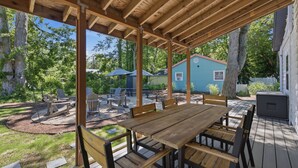 An outdoor patio with wooden tables and chairs, a covered seating area, and a view of a blue house in the background. - Union Pier Cottage Close To Everything! One Of Three Units! (Union Pier)