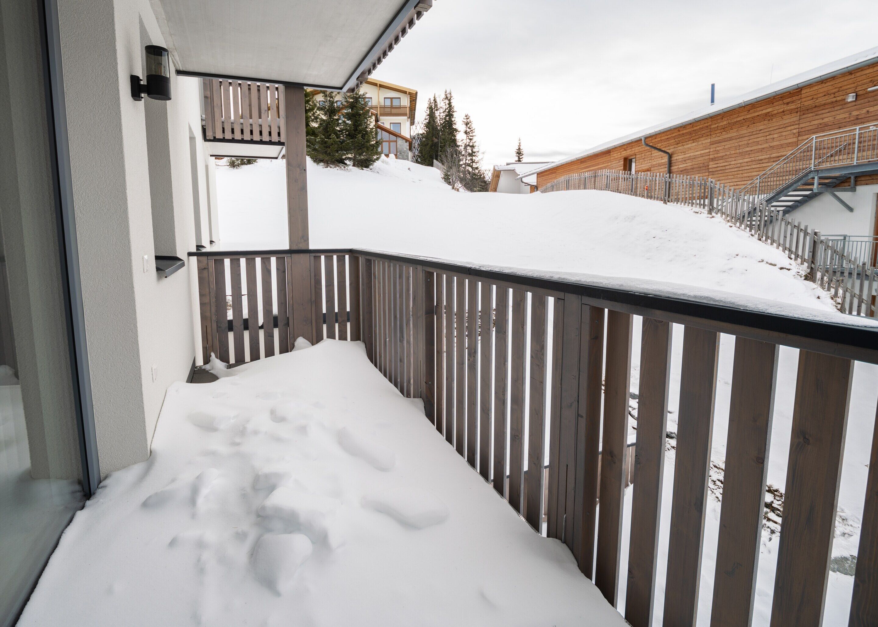 A snow-covered balcony with a wooden railing, a view of a building with a wooden facade, and a staircase leading up to a wooden deck.