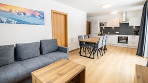 A modern apartment with a living area, dining table, grey sofa, wooden coffee table, kitchen with white cabinets, and a painting of a mountain landscape. - Apartment in Katschberghohe Near Ski Slopes (Rennweg am Katschberg)