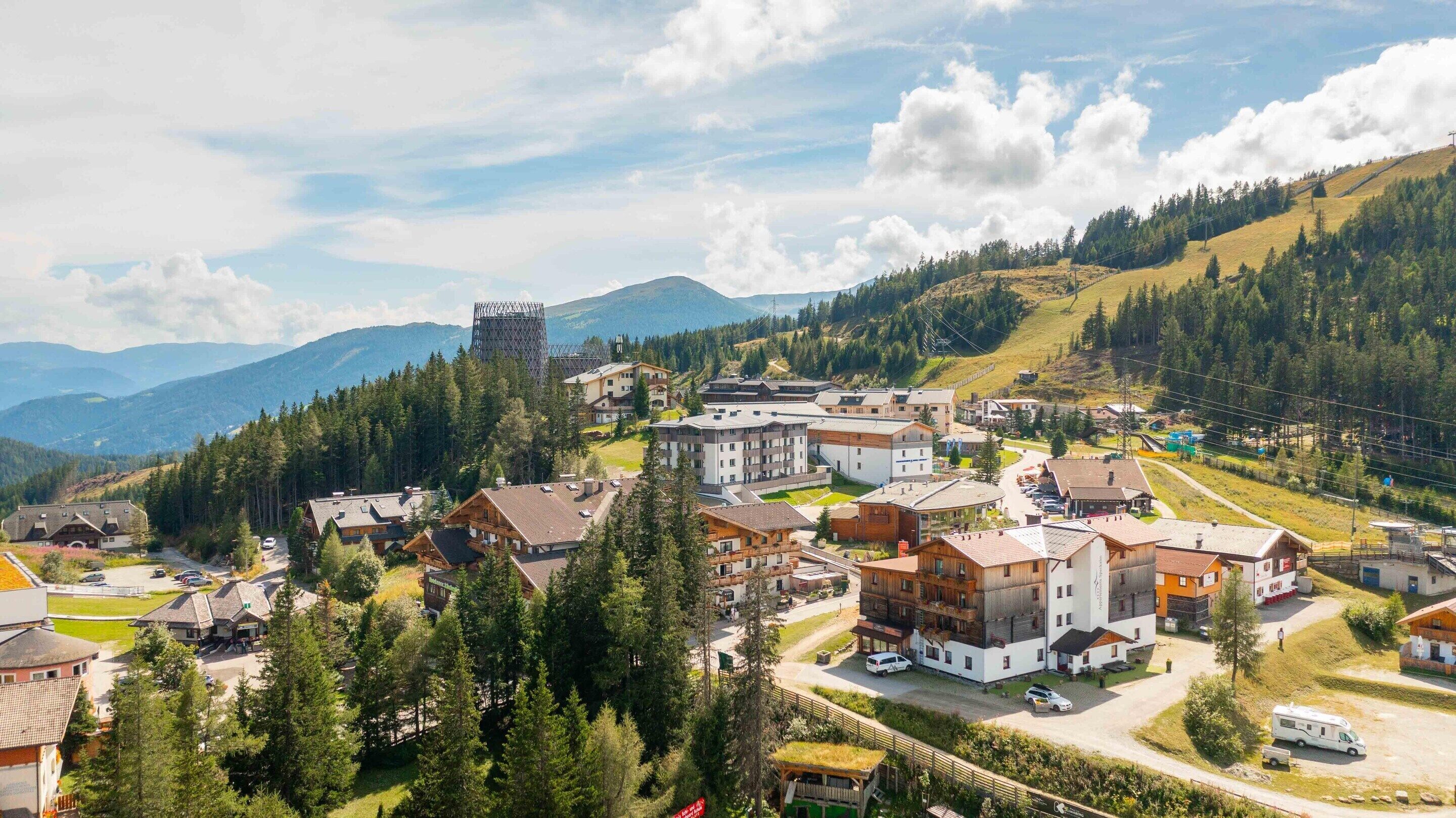 Ein Bergdorf mit traditionellen Holzhäusern, einer Seilbahn und Blick auf die umliegenden Berge.