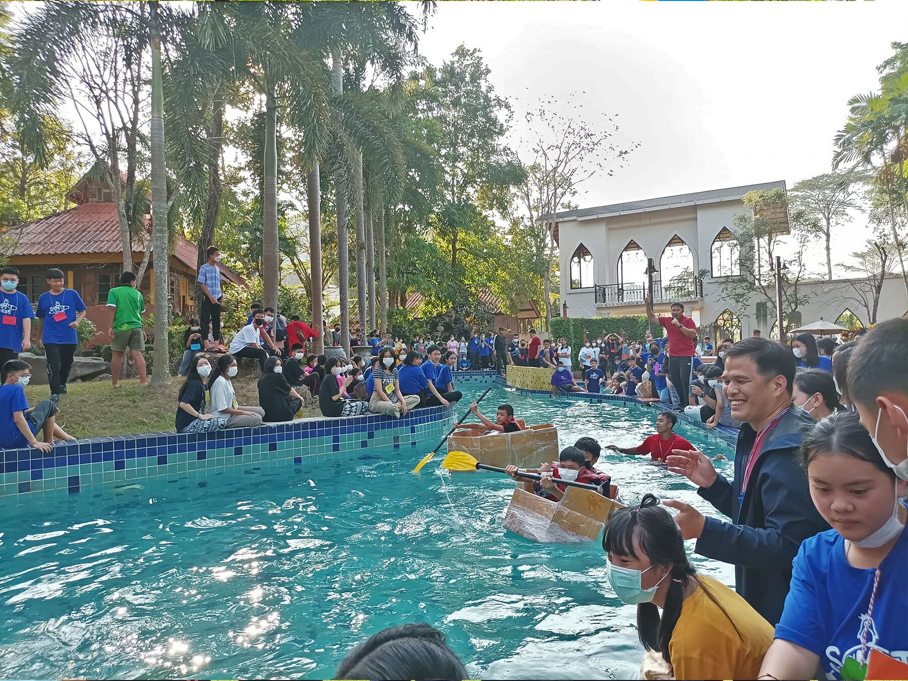 A swimming pool with people in boats and on the edge, surrounded by trees and a building with arched windows.