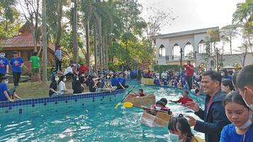 Una piscina con gente en botes y en los bordes, rodeada de árboles y un edificio con ventanas arqueadas.