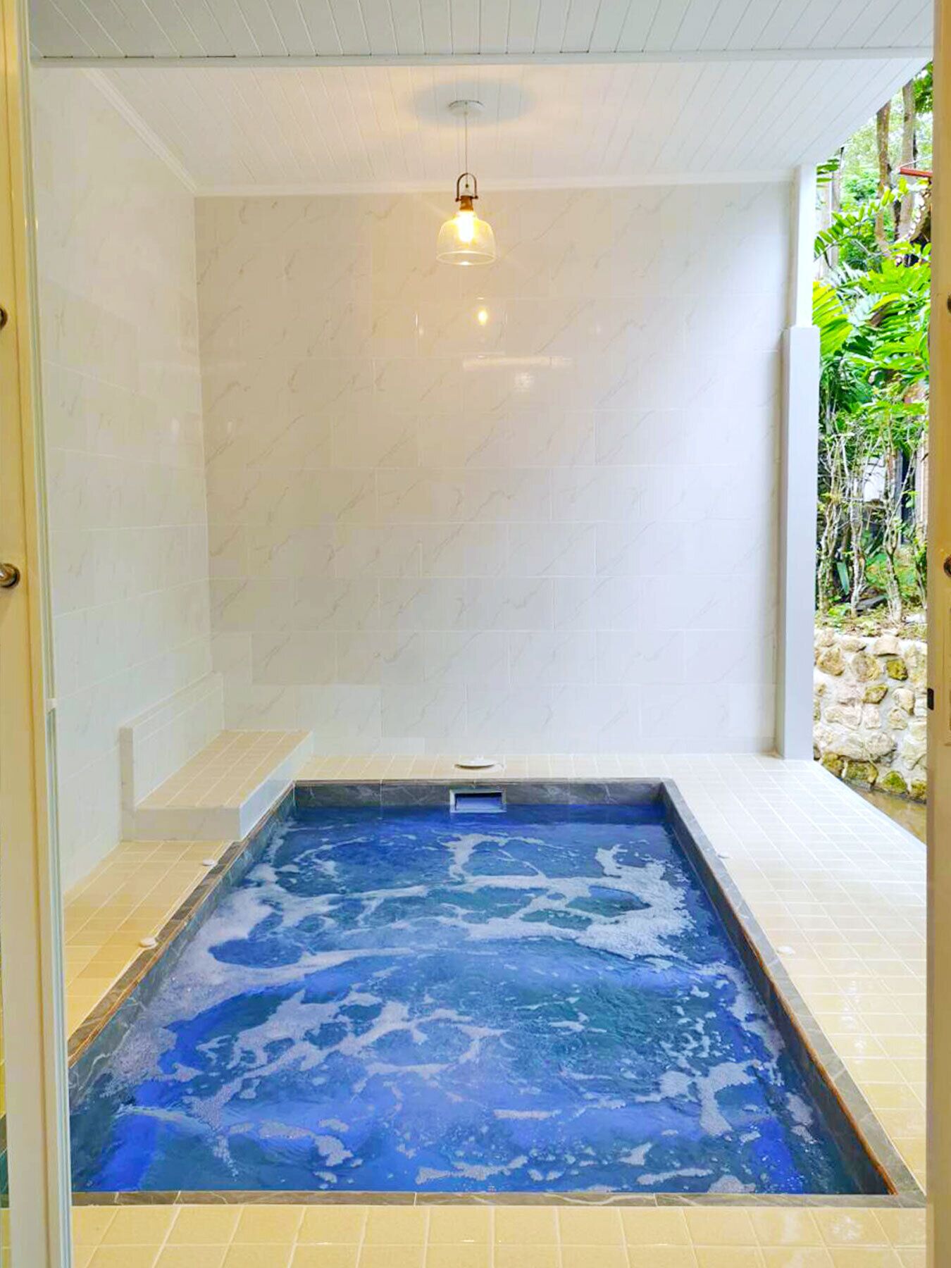 A spa area with a hot tub, tiled walls, and a stone wall with greenery visible through an open door.