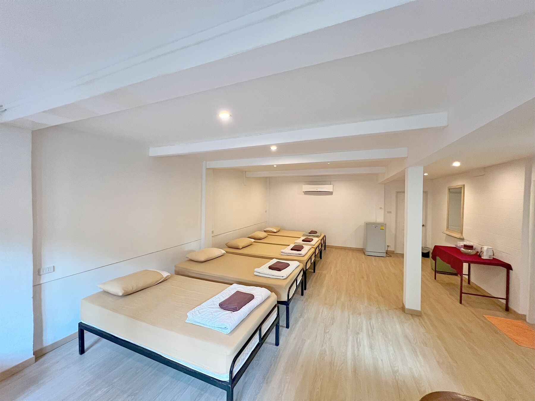 A hotel room with multiple beds aligned in a row, a small table with a red tablecloth, and a mirror on the wall.