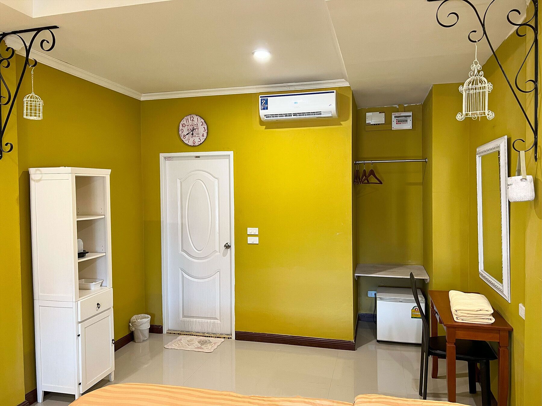 A room with a white door, a white wardrobe, a small table with a towel, a mirror, and a clock on the wall.