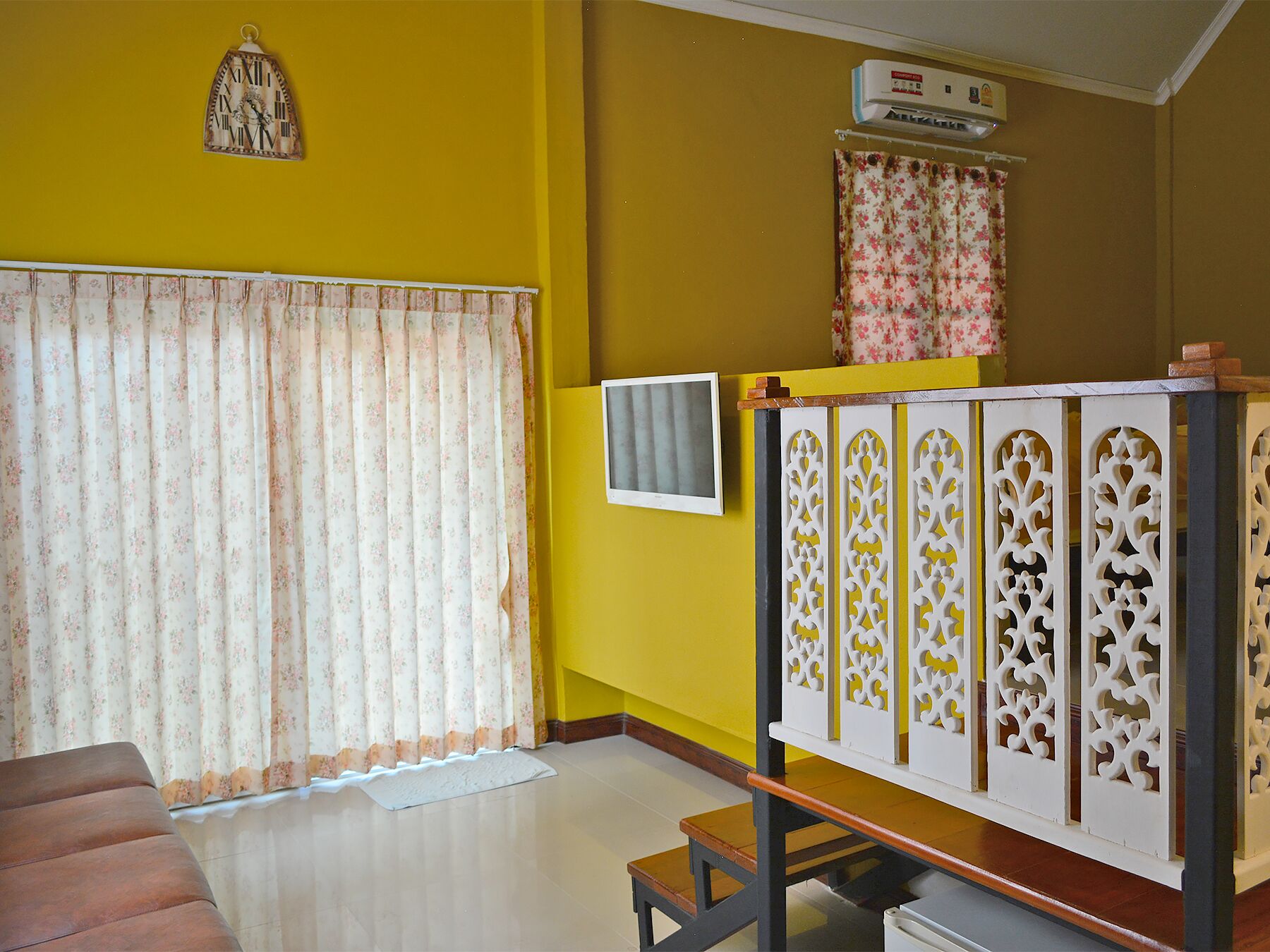 Una habitación con paredes amarillas, una cortina estampada con motivos florales, un separador decorativo blanco, una mesita y un reloj fijado a la pared.