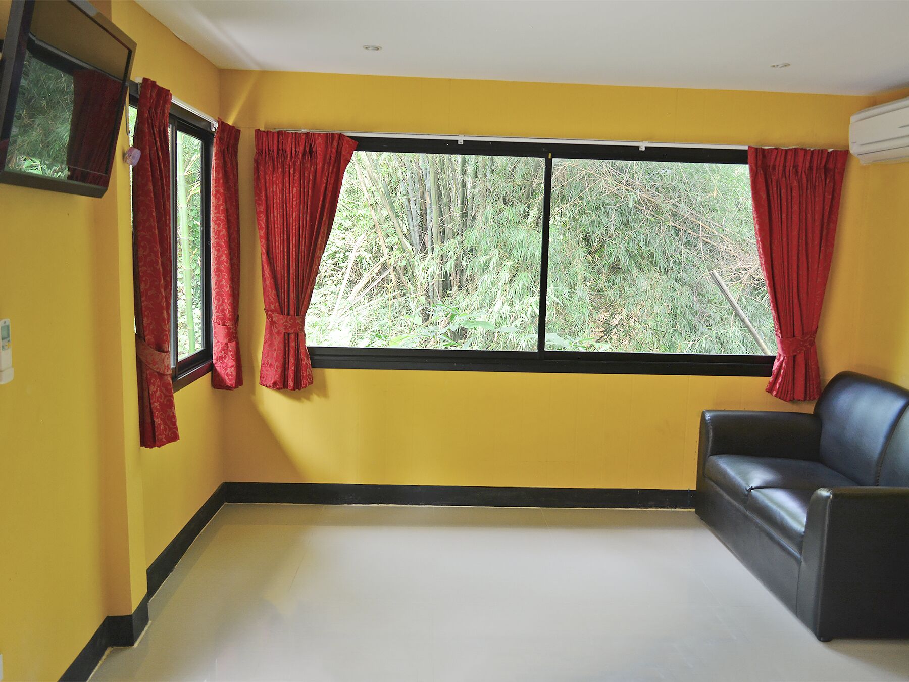 Una habitación con paredes amarillas, un sofá de cuero negro y un ventanal con cortinas rojas.