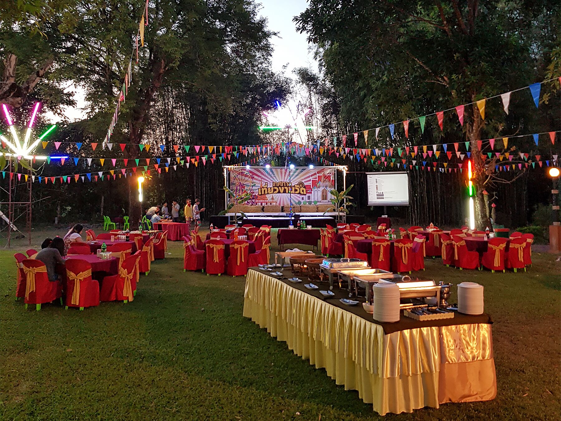 An outdoor event with tables and chairs arranged for guests, a stage with a banner, and colorful decorations.