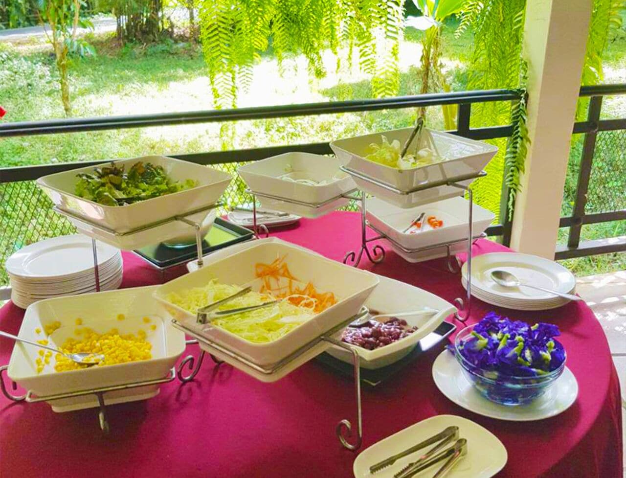 Een buffetstafel met diverse gerechten en bestek, buiten opgesteld met uitzicht op groen.