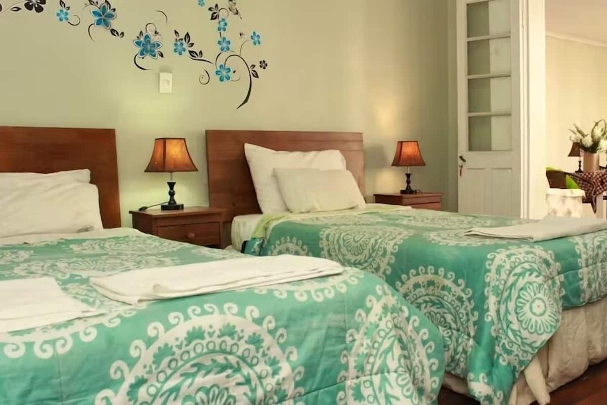 Quarto de hotel com duas camas, cada uma com colcha verde estampada e almofadas brancas. Há duas luminárias de mesa, uma cabeceira de madeira e uma decoração na parede com flores azuis e pretas.