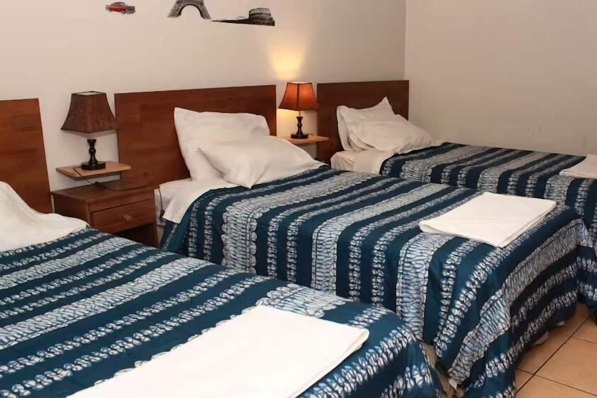 Quarto de hotel com duas camas de solteiro, cada uma com cobertura estampada azul e branca, almofadas brancas e uma pequena mesa de cabeceira com abajur.