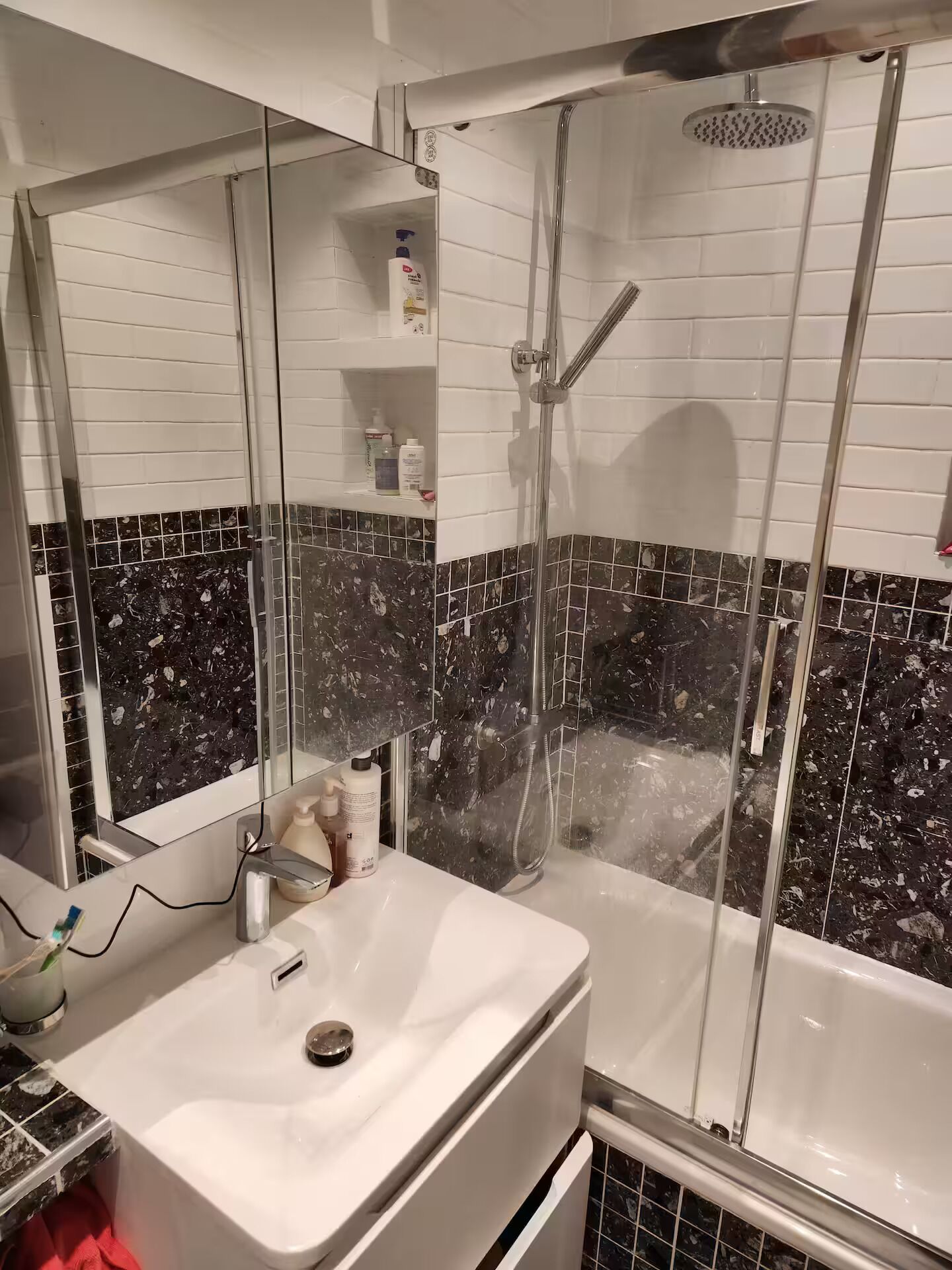 Een badkamer met een witte wastafel, een spiegel, een douchecabine en een betegelde muur met een marmerachtig patroon.