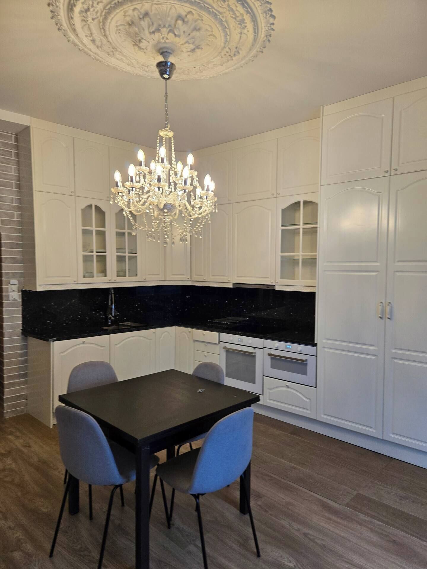 Une cuisine avec des meubles blancs, un plan de travail noir, un lustre et une table à manger avec des chaises.