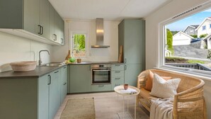 A modern kitchen with light green cabinets, a central island, and a built-in oven. There's a wicker sofa with cushions by a window overlooking a residential area. - Delightful 2-bedroom apartment in charming Lillehammer (Innlandet)