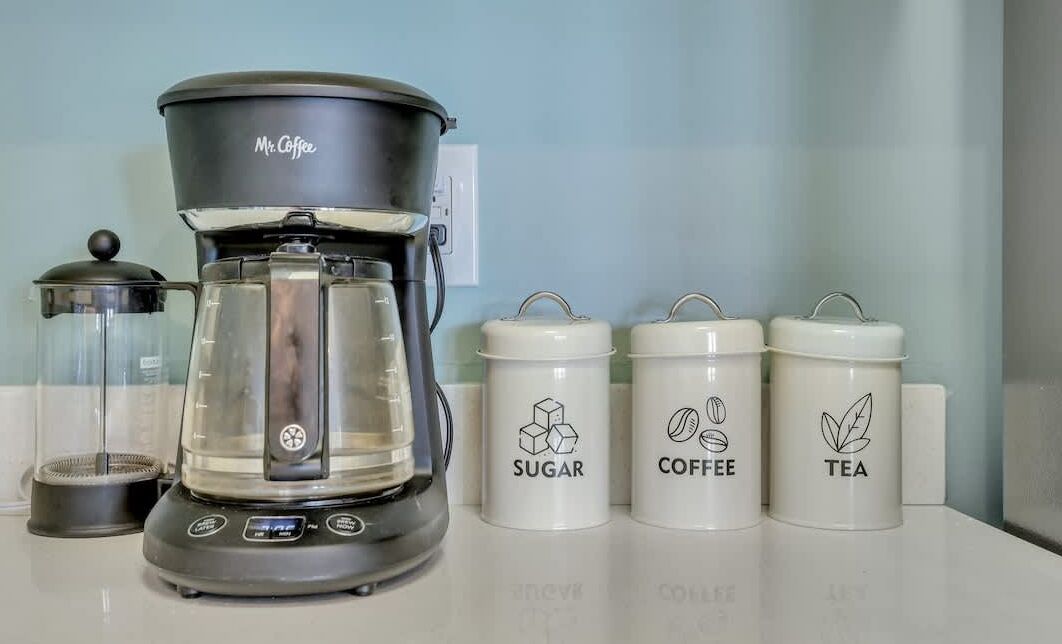 A coffee maker labeled 'Mr. Coffee' with three containers labeled 'SUGAR,' 'COFFEE,' and 'TEA' on a countertop.