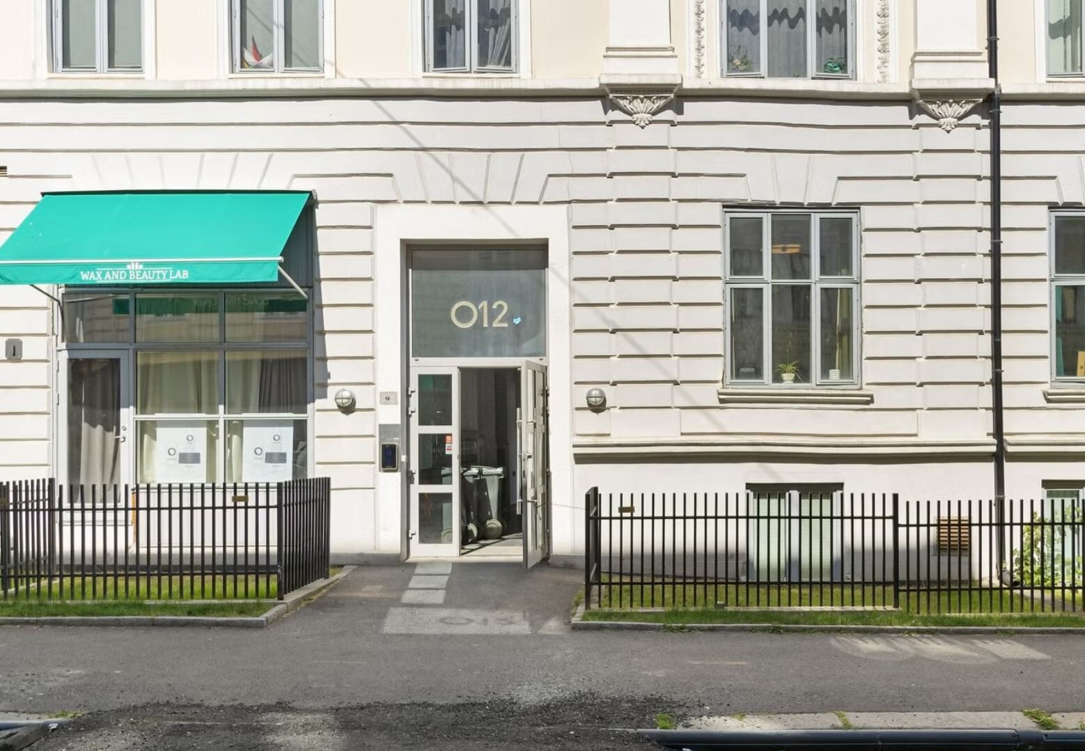 Ein Gebäude mit einer grünen Markise, auf der 'WAX AND BEAUTY LAB' steht, und einer Tür mit der Aufschrift 'O12'.