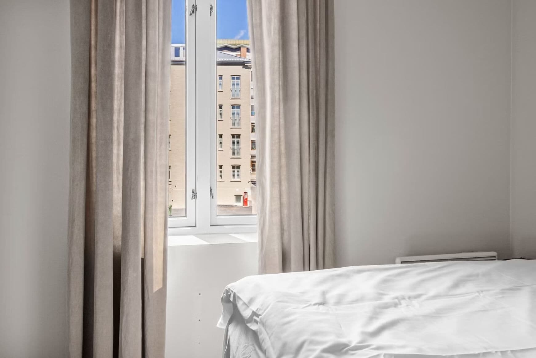 A bedroom with a bed, a window with curtains, and a view of buildings outside.