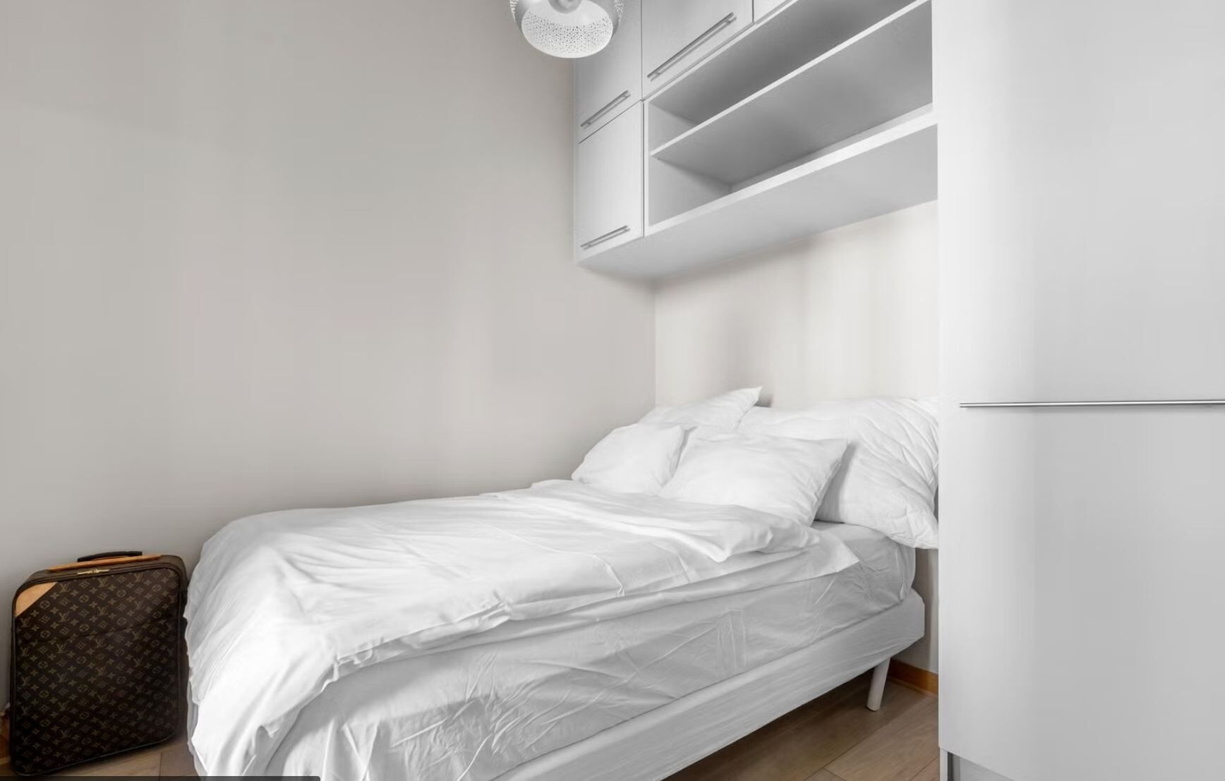 A single bed with white bedding, a suitcase, and built-in storage cabinets above the bed.
