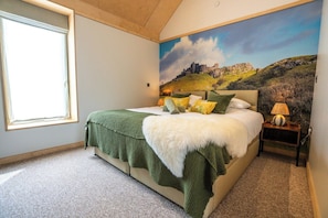 A bedroom with a large bed, a window, a painting of a landscape, and a nightstand with a lamp.