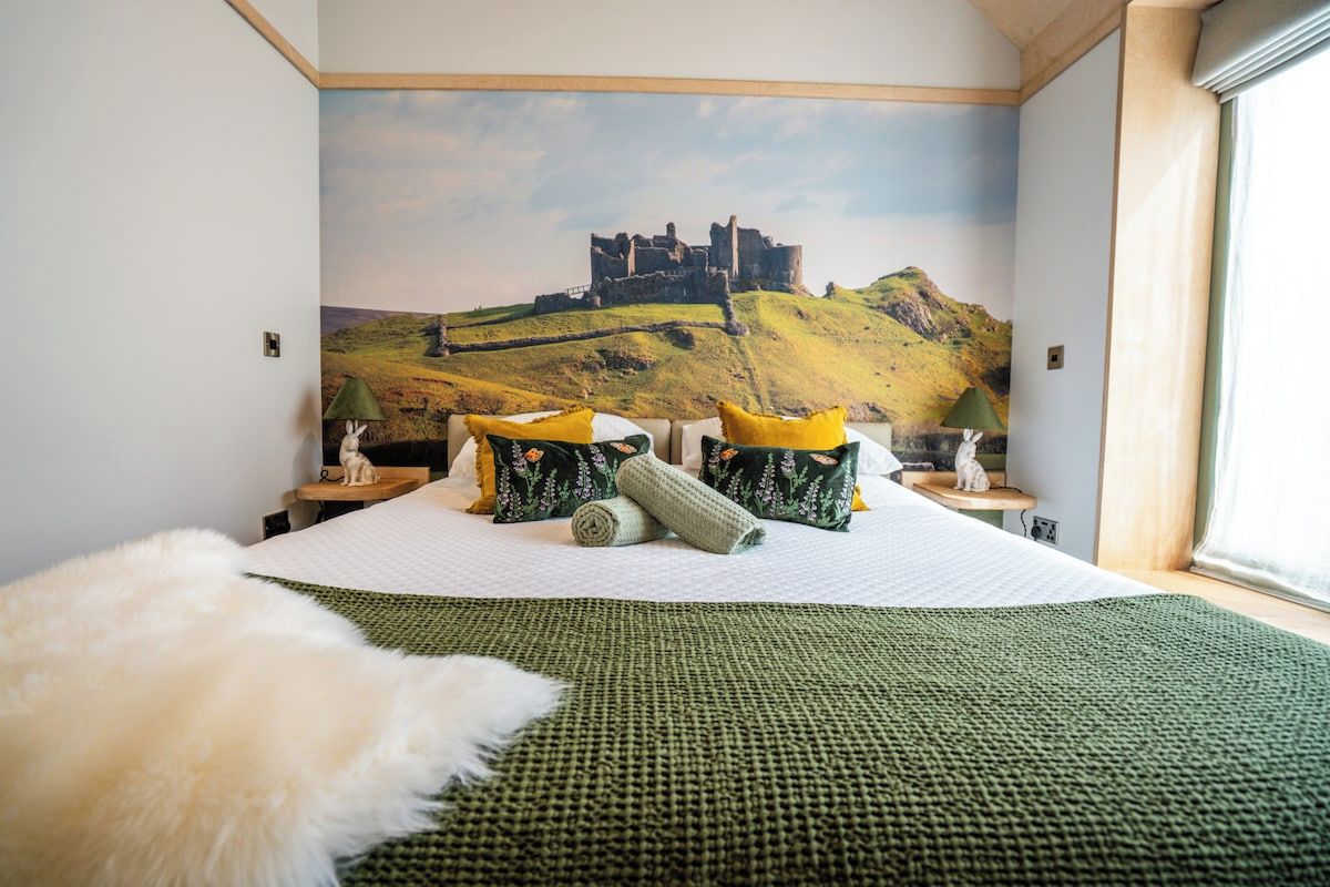 A bedroom with a large bed, a green knitted blanket, and a wall mural of a castle on a hill.