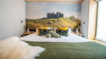 A bedroom with a large bed, a green knitted blanket, and a wall mural of a castle on a hill.