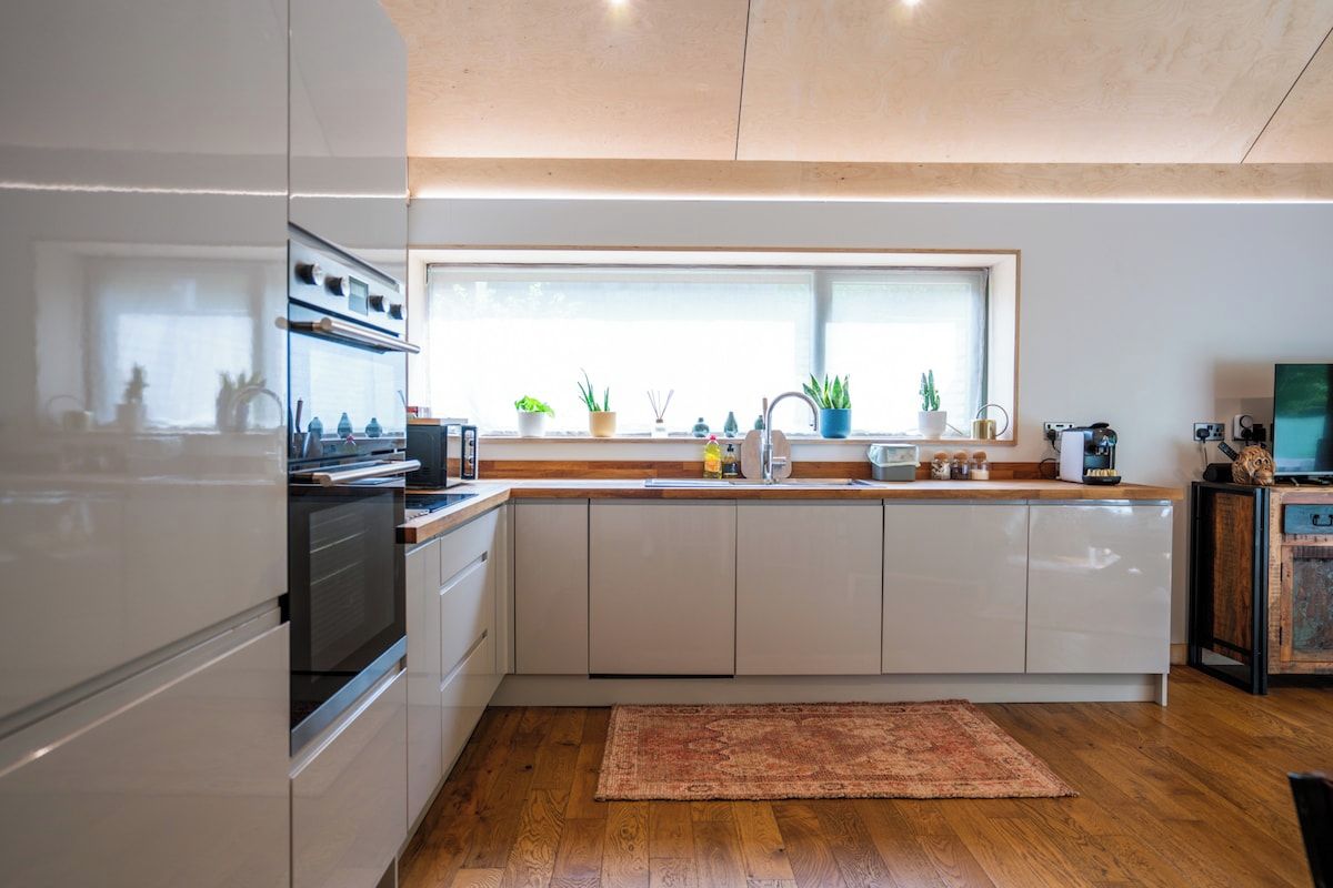 Una cocina moderna con armarios blancos, horno empotrado, ventana grande y suelo de madera.