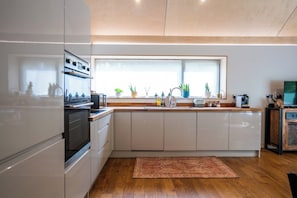 A modern kitchen with white cabinets, a built-in oven, a large window, and a wooden floor.