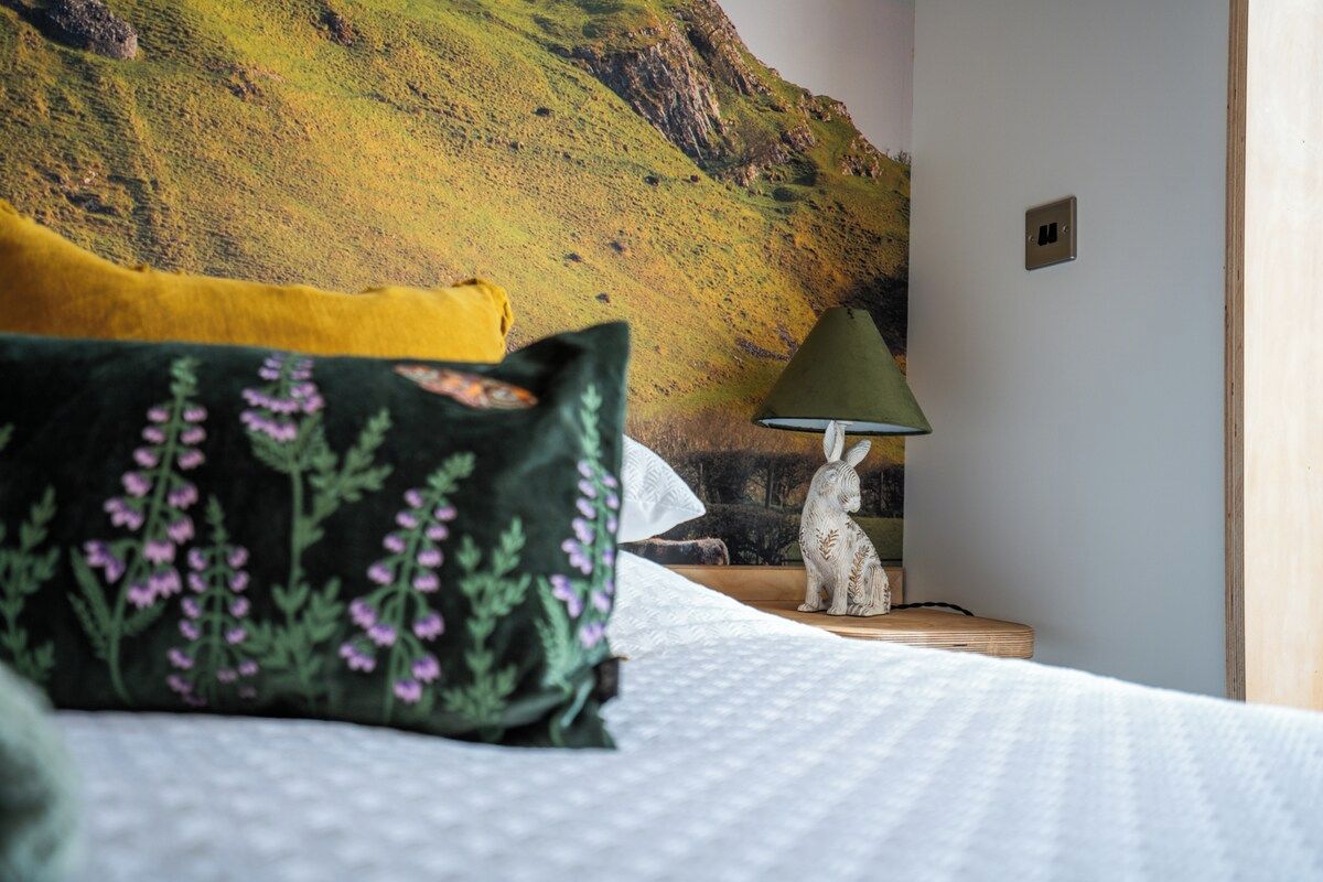 A bed with a white duvet, a green pillow with floral patterns, a yellow pillow, and a white rabbit figurine beside a lamp on a bedside table.