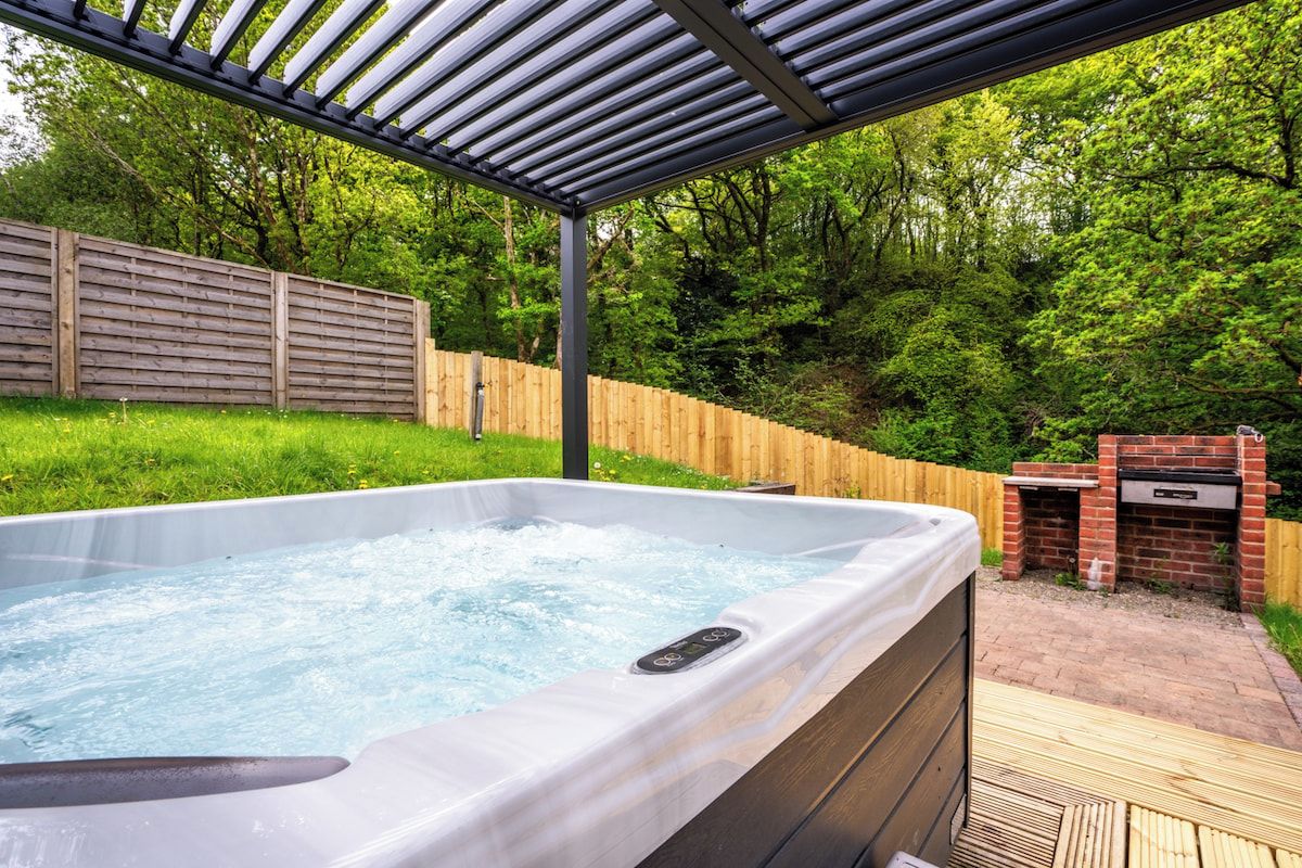 A hot tub under a pergola with a wooden fence and a brick barbecue in the background.