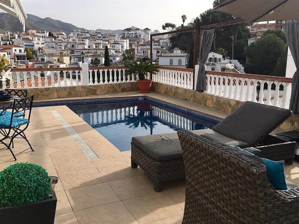 A rooftop pool area with a view of a coastal town, including a pool, lounge chairs, and a potted plant.