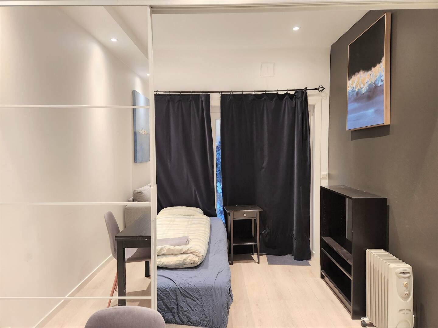 Et moderne soveværelse med seng, skrivebord, stol, radiator og et billede på væggen.