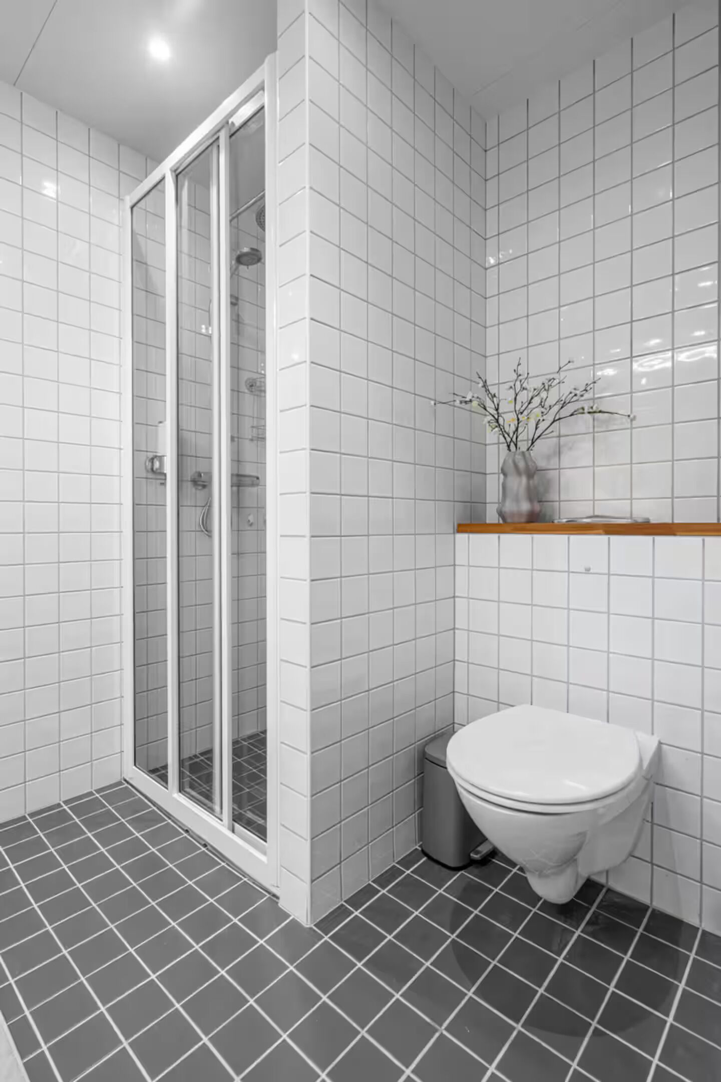 A modern bathroom with white tiles, a glass shower enclosure, a white toilet, and a small shelf with a vase.