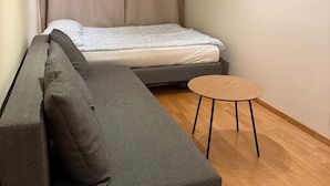 Una stanza compatta con un divano grigio, un piccolo tavolino rotondo in legno e un letto con copriletto bianco e grigio.