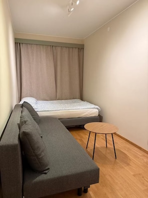 Ett litet rum med en grå soffa, ett litet runt träbord och en säng med ett vitt och grått mönstrat överkast.