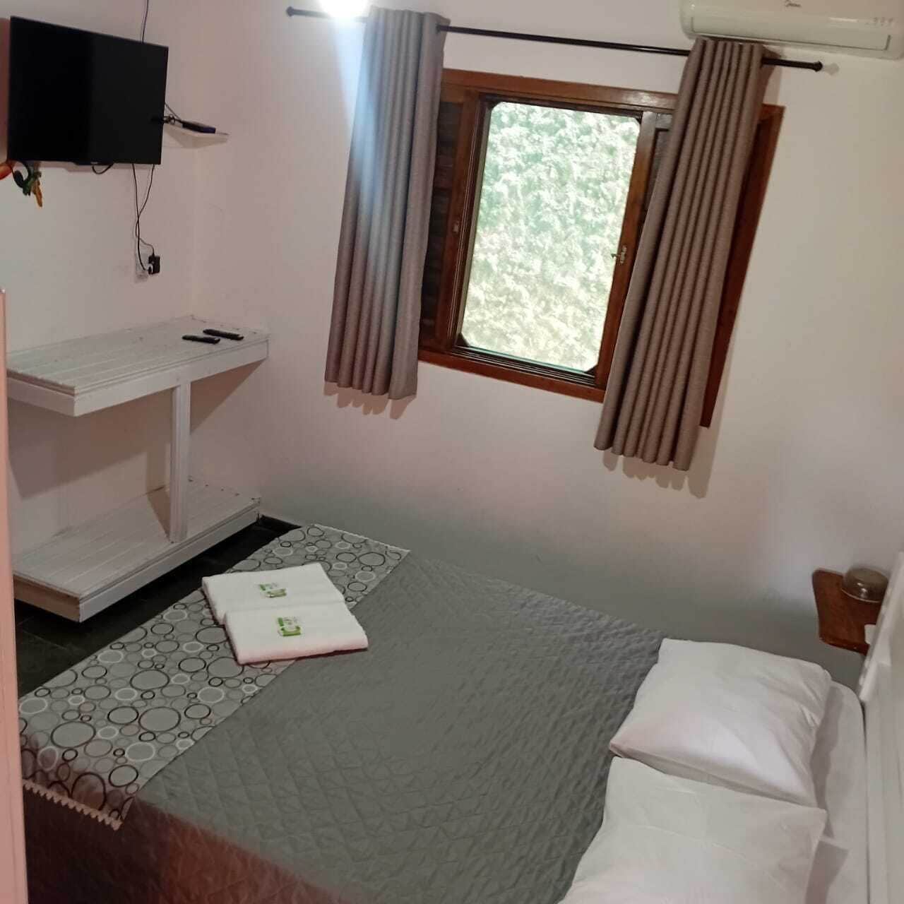 Quarto pequeno e simples com uma cama, uma estante branca, uma janela com cortinas e uma TV fixada na parede.