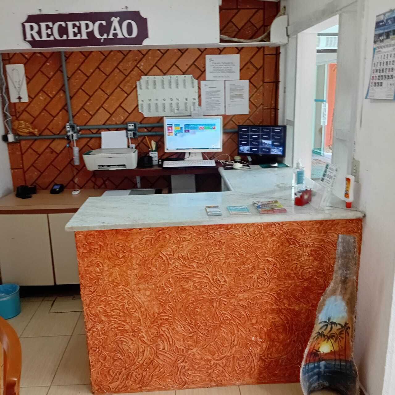 A reception area with a sign reading "RECEPCAO," a computer, a printer, and a counter with a patterned orange design.