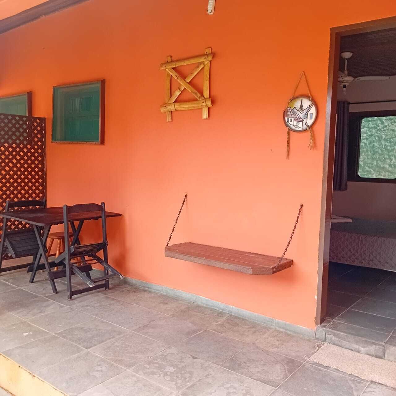 Quarto com parede laranja, balanço de madeira, mesa pequena com cadeiras e uma cama ao fundo.