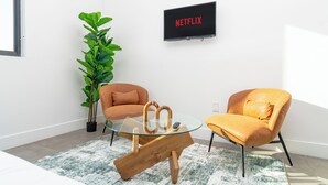 Een moderne woonkamer met een glazen salontafel, twee leren fauteuils, een grote groene plant en een televisie waarop 'NETFLIX' staat.