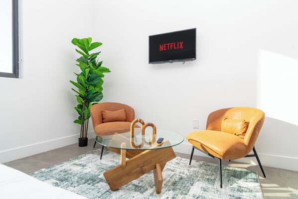 Een moderne woonkamer met een glazen salontafel, twee leren fauteuils, een grote groene plant en een televisie waarop 'NETFLIX' staat.
