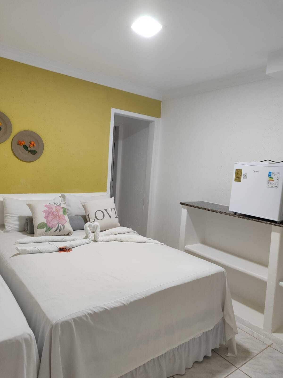 A hotel room with a bed, a small table with a refrigerator, and a yellow accent wall.